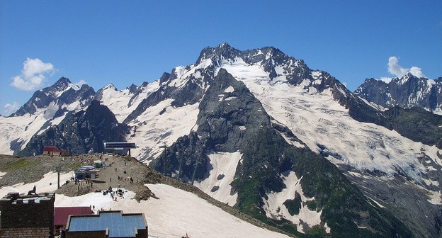 Вид с горы Орлиное гнездо на Дамбай. Фото LxAndrew https://ru.wikipedia.org/wiki/Домбай_(горная_территория)#/media/Файл:62_ГКХ,_г._Джугутурлючат,_Инэ,_верхняя_станция.JPG